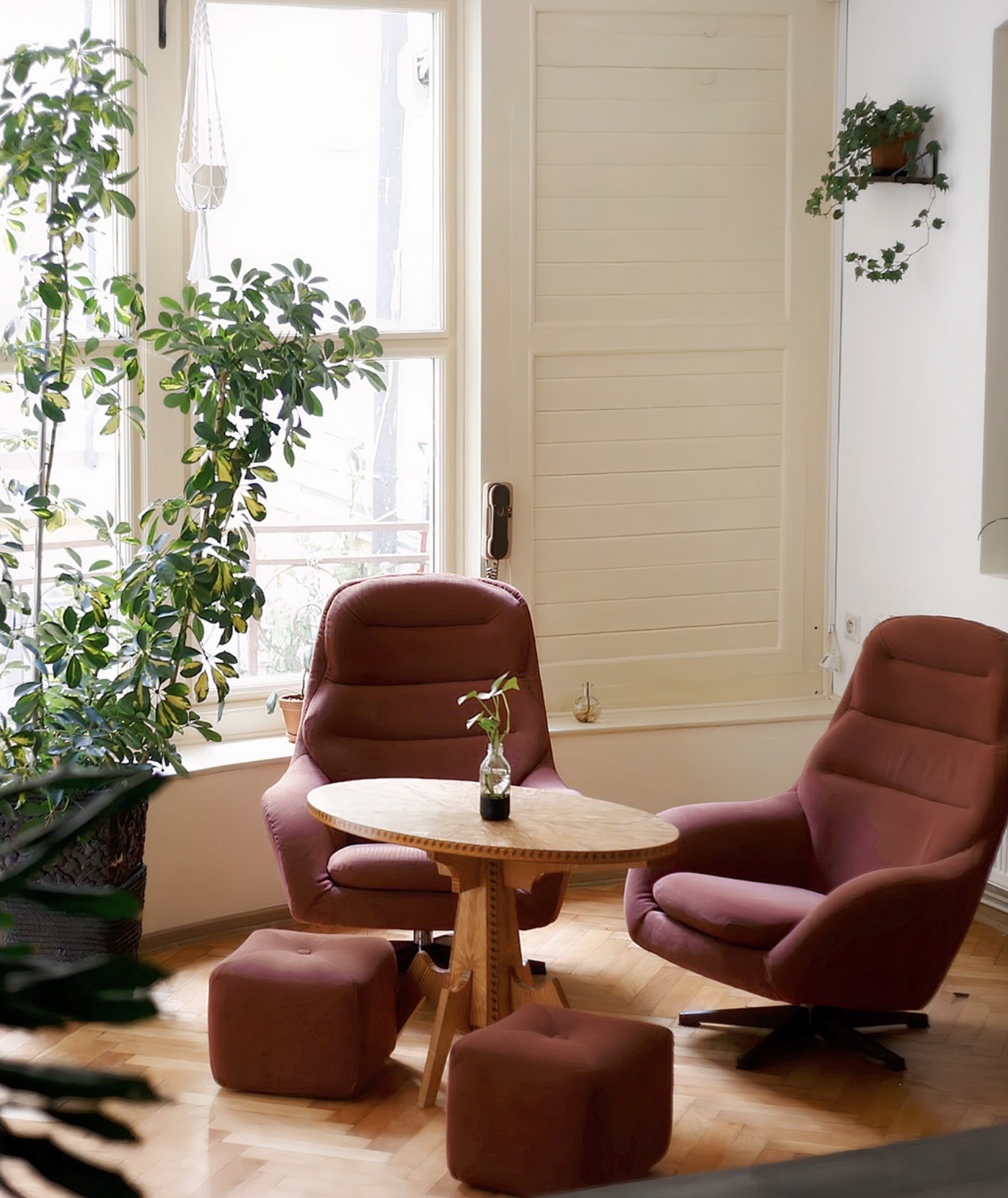 Two vintage rust colored armchairs with matching ottomans around a small round table, sunlit by tall windows and surrounded by houseplants